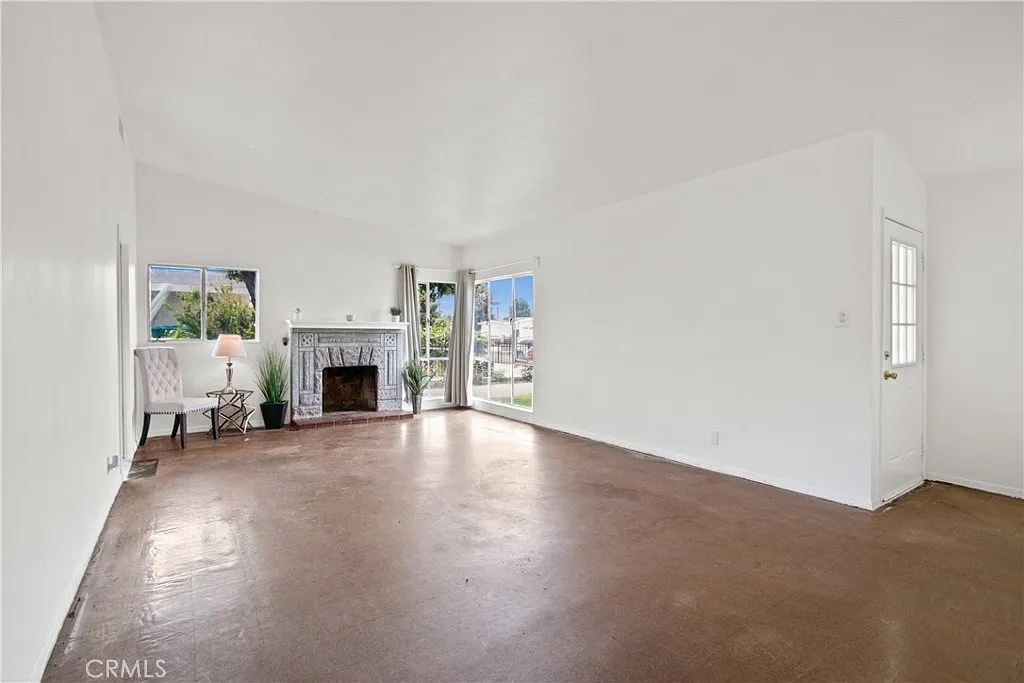 living room with white walls, bare floor, fireplace, large windows, a door outside, and a chair by the fireplace.