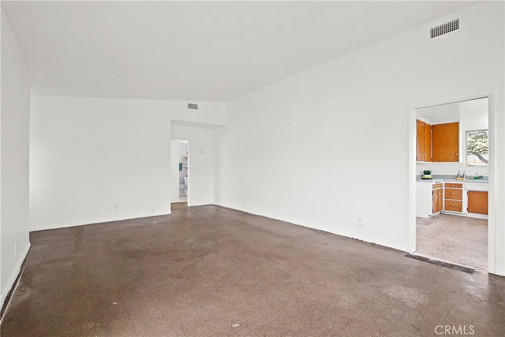 Empty room with white walls, concrete floor, and a doorway showing a kitchen with wooden cabinets.