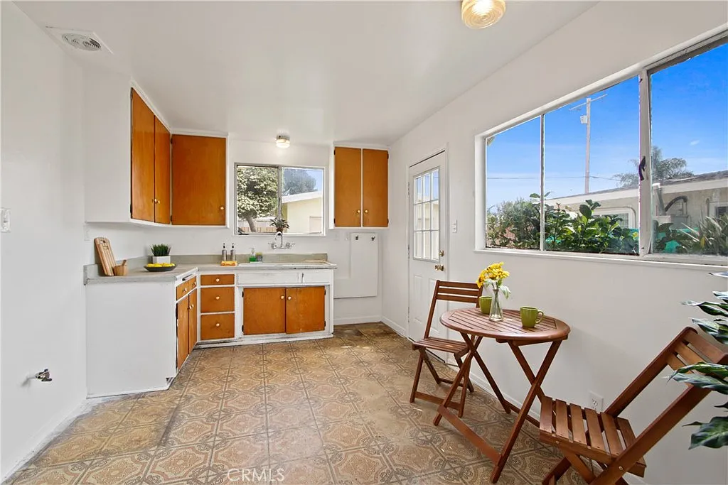 Kitchen with white walls, wooden cabinets, a window, a small table with flowers, two wooden chairs, and patterned floor.