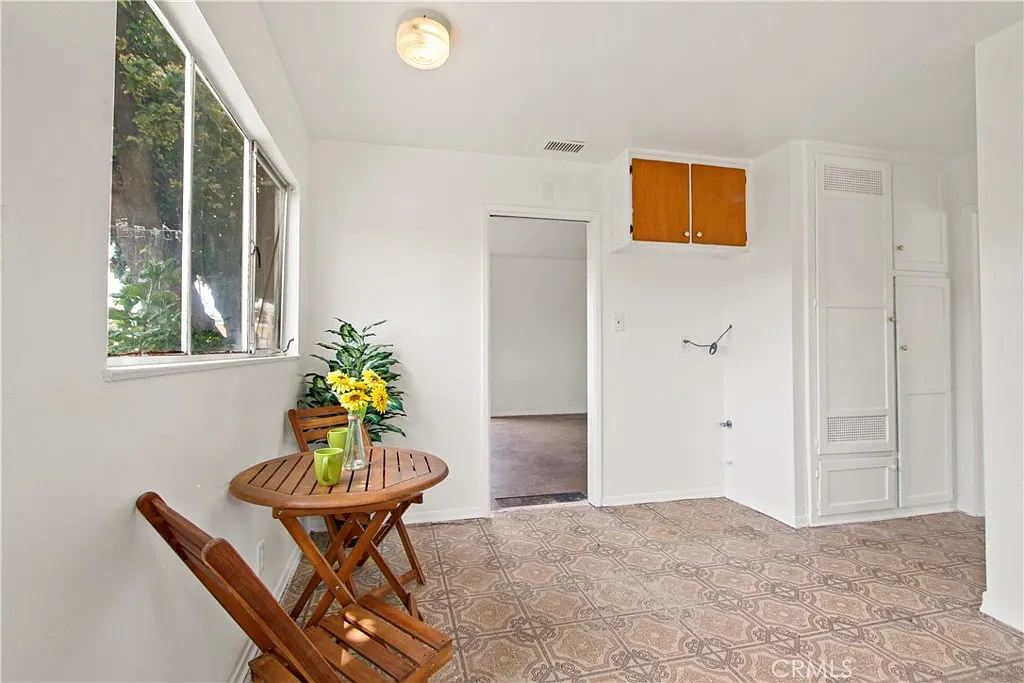 A bright room with a small table, two wooden chairs, yellow flowers, a window, and patterned tile floor.