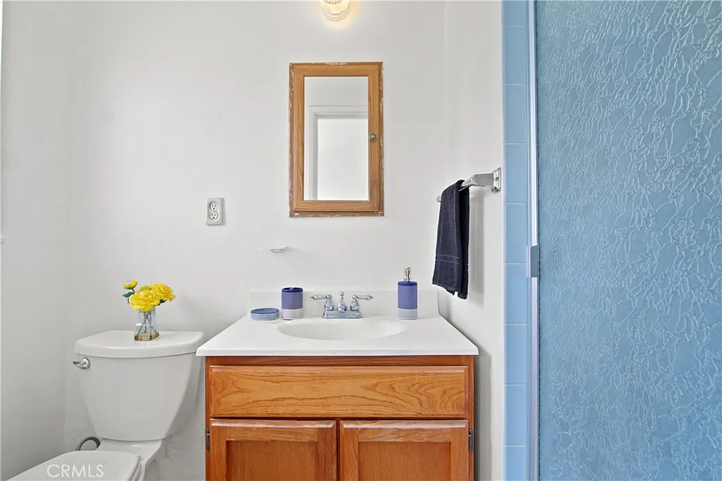 A small bathroom with white toilet, wooden sink cabinet, mirror, frosted glass shower, and yellow flowers on the counter.