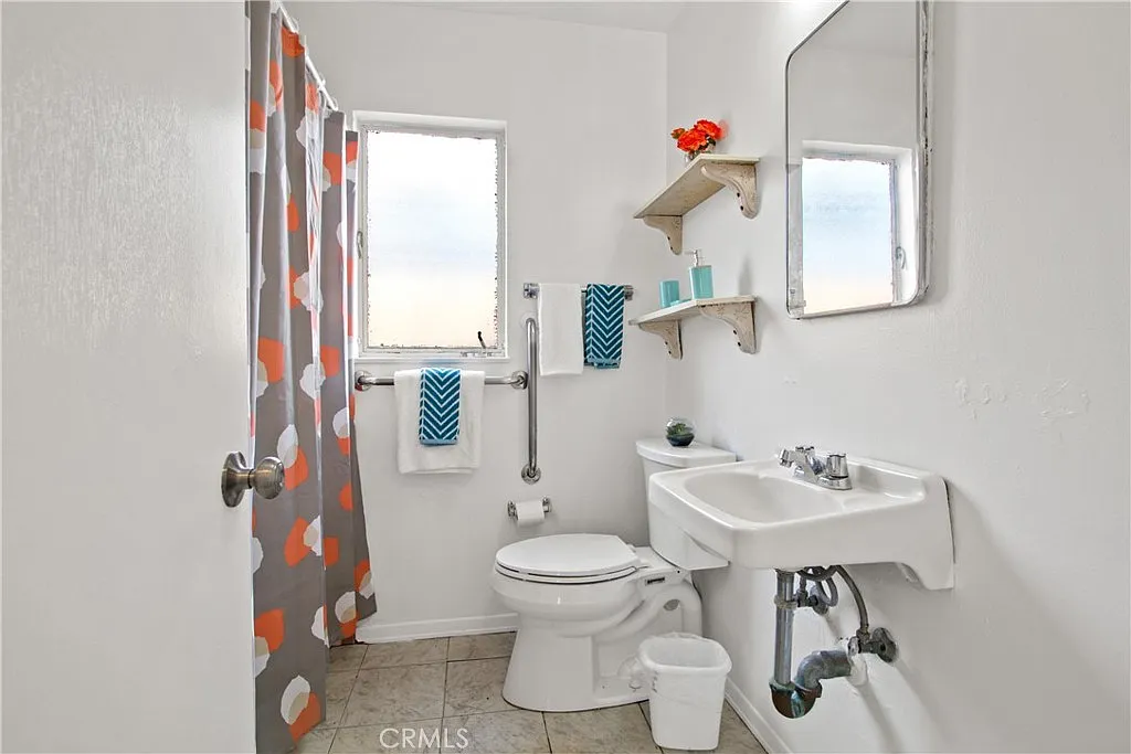 Bathroom with white sink and toilet, patterned shower curtain, shelves with decor, and sunlight from a window.