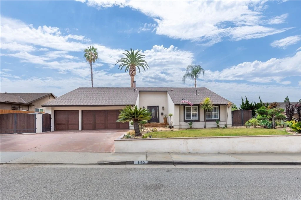 Single-story real estate house with landscaped front yard, palm trees, and two-car garage, light-colored exterior