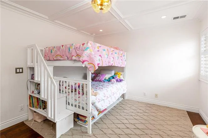 Children's bedroom with white bunk bed, colorful unicorn blanket, shelves with books and toys, neutral walls, and soft lighting