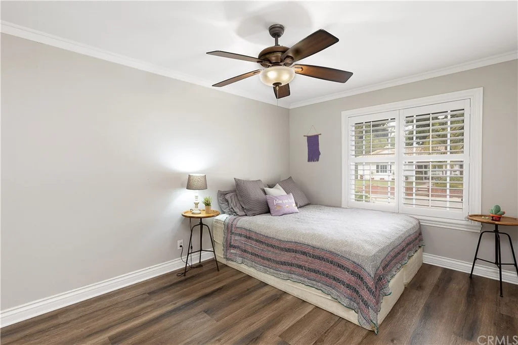 Bedroom with bed, gray bedding, two side tables with lamps, ceiling fan, window shutters, and light brown wooden floor