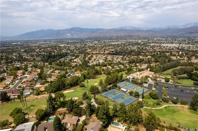 Aerial view of suburban area with houses, green spaces, tennis courts, and mountains in background
