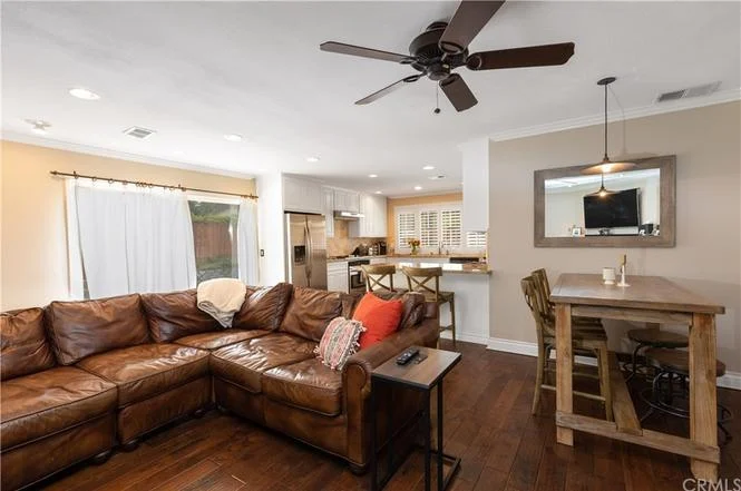 living room with brown leather sectional sofa, side table, dining area, ceiling fan, light walls, large windows with white curtains, and kitchen with modern appliances