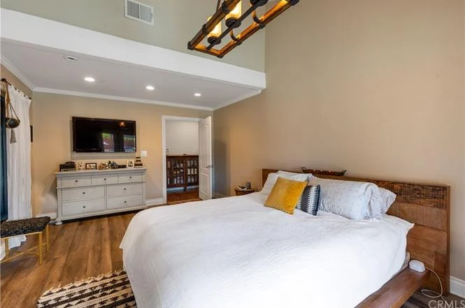bedroom with large bed, white bedding, yellow pillow, dresser, wall-mounted TV, wooden floor, and doorway to another room