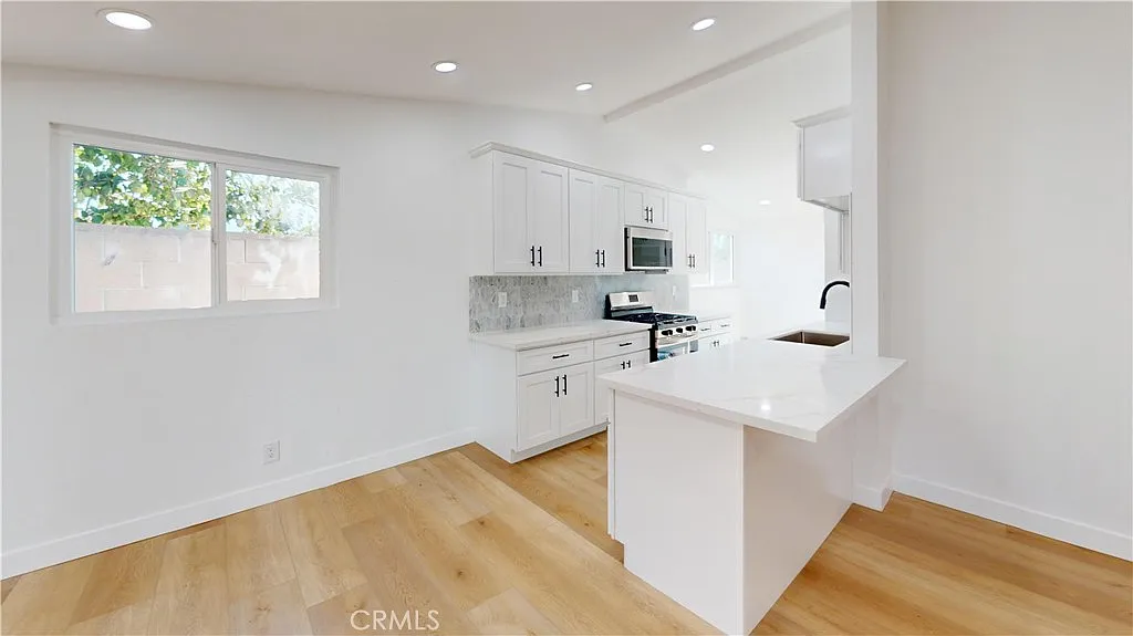 A kitchen with white cabinets, stainless steel microwave, island with sink, and light wood floors with sunlight.