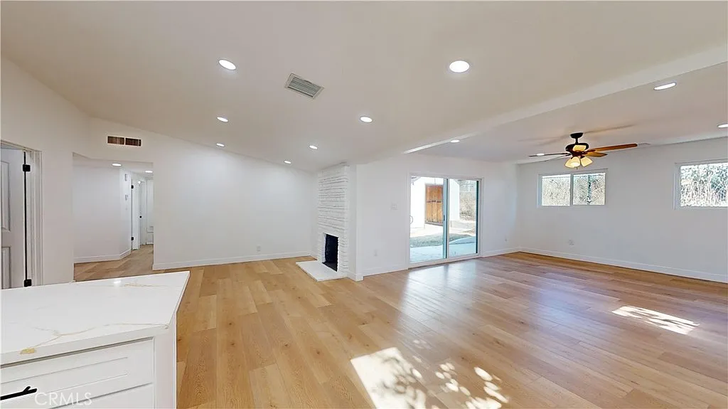 A bright living room with light wood floors, white walls, a ceiling fan, and a fireplace, with windows letting in sunlight and doors leading outside.