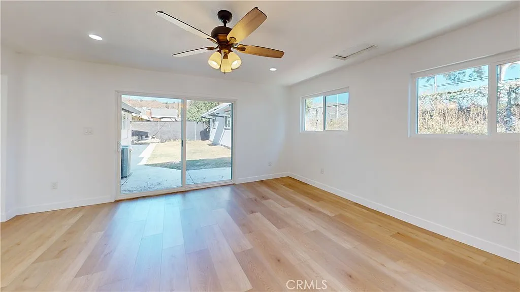 Empty room with light wood floor, ceiling fan, large windows, and sliding glass door to outside.