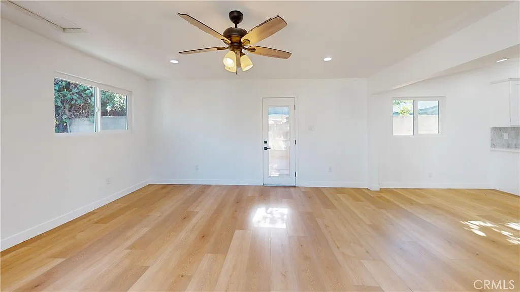 Empty living room with light wood floor, ceiling fan, large windows, and door to outside.