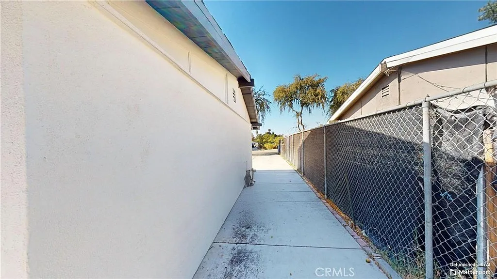 Narrow pathway between buildings with concrete surface, fence, and trees in background.