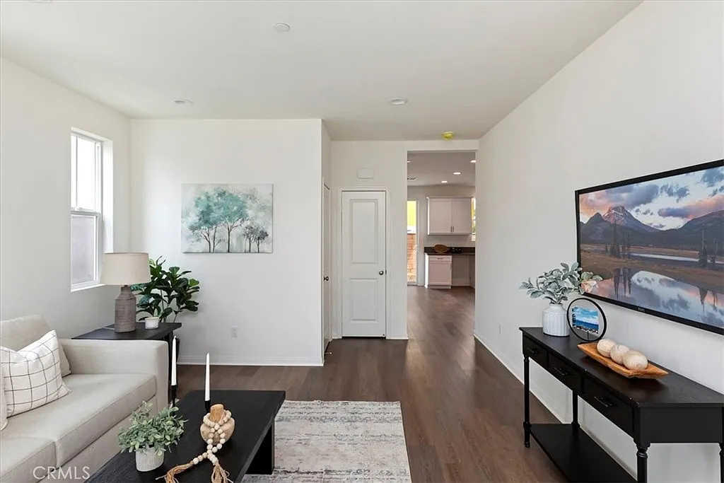 Living room with light sofa, plants, TV on black stand, white walls, hardwood floor, and door to another room.