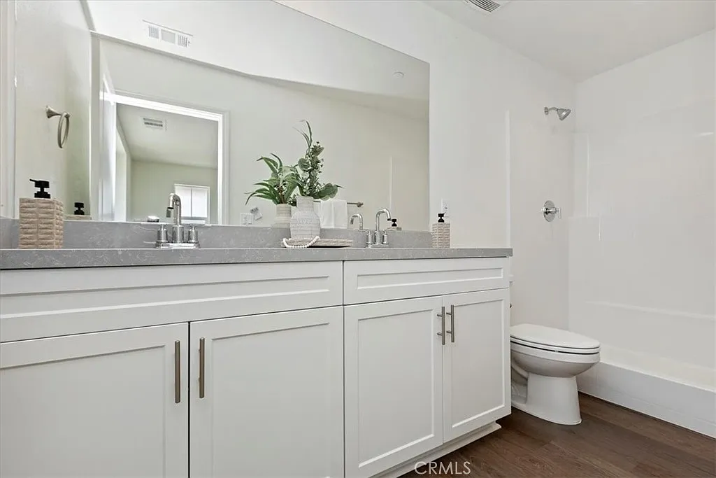 Bathroom with double sink vanity, gray countertops, large mirror, shower, and minimal decor with plants and soap dispensers.