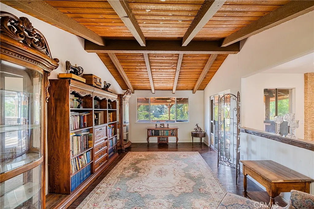 Room with wooden beam ceilings, bookshelves, glass cabinet, table, and large windows