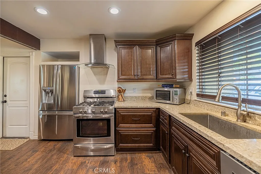 Kitchen with dark wood cabinets, stainless steel appliances, granite countertops, gas stove, microwave, and sink by window with blinds.