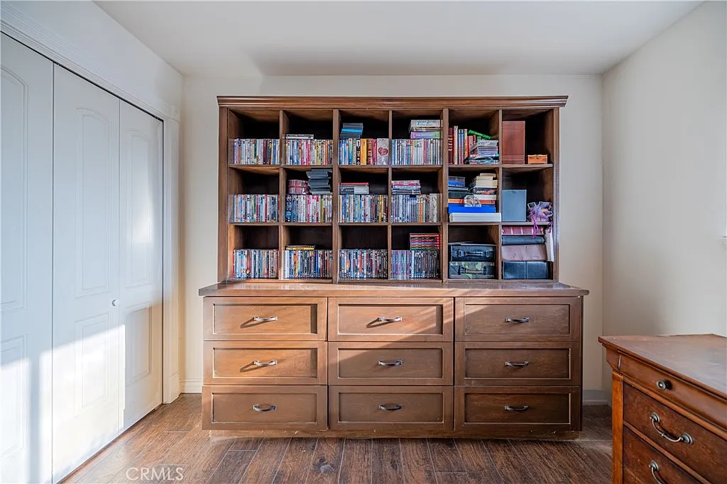 Wooden dresser with drawers, bookshelf filled with DVDs, and two closed closet doors in the background.