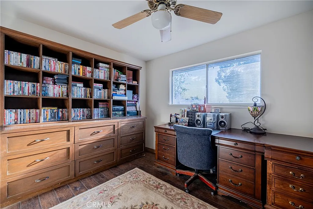 Home office with wooden desk, chair, large window, built-in shelves with books and dvds, and decorative rug.