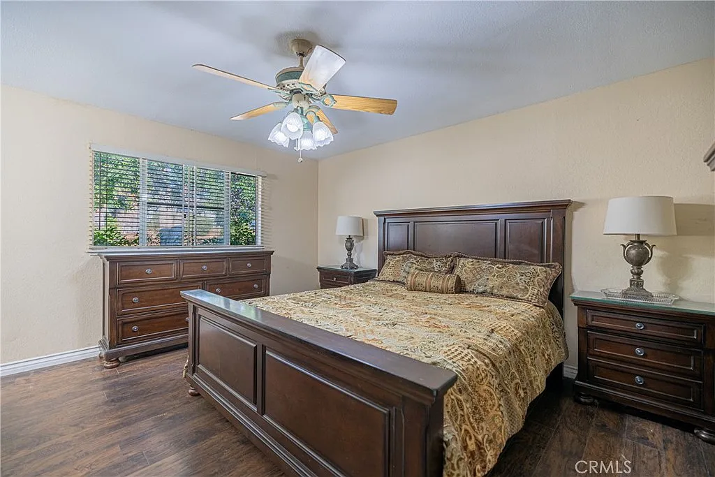 Bedroom with wooden bed, patterned bedspread, matching nightstands with lamps, dresser, ceiling fan, and windows with blinds.