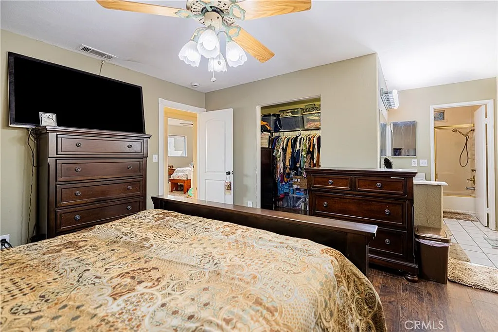 Bedroom with wooden bed, patterned blanket, dresser with tv, open closet with clothes, and adjacent bathroom.