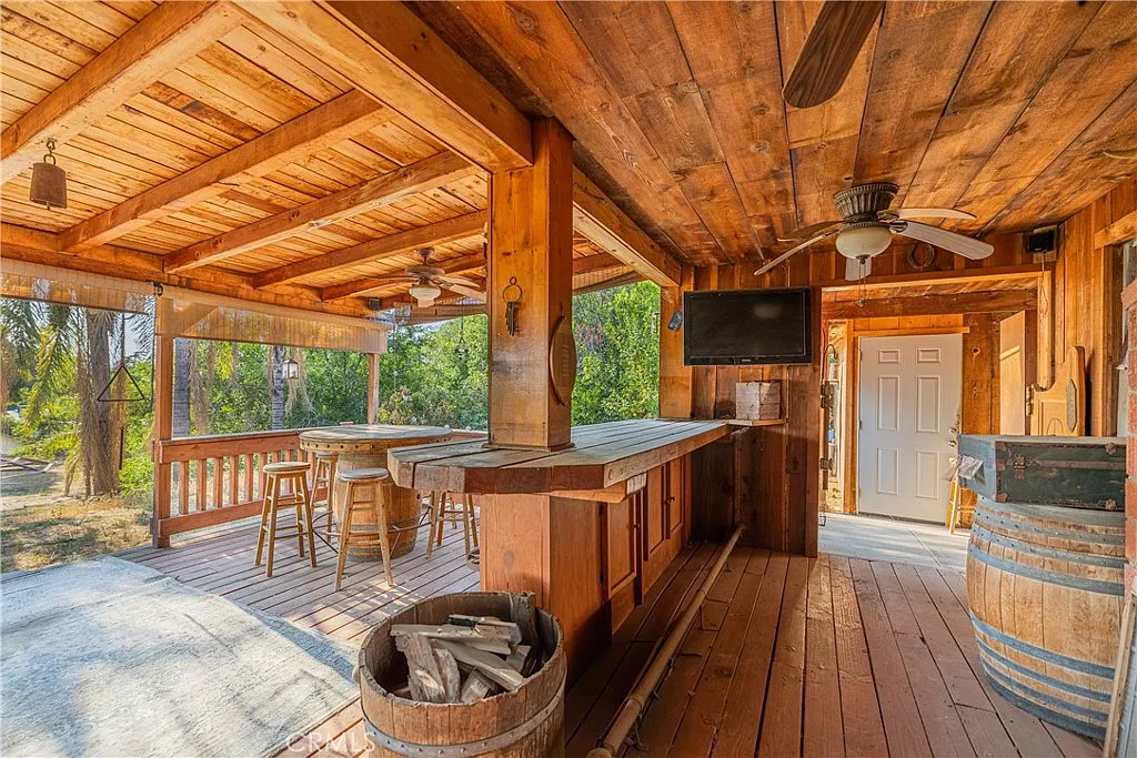 Wood porch with bar, stools, wall TV, barrels, and green plants.
