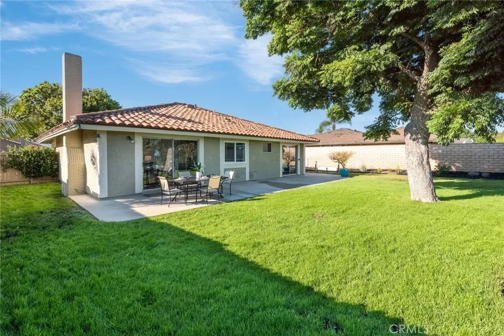 Single-story house real estate with tile roof, patio seating, grass, and trees.