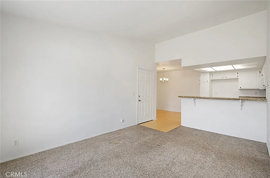 Living space with light walls, carpeted floor, and adjacent kitchen or dining area with bar counter.
