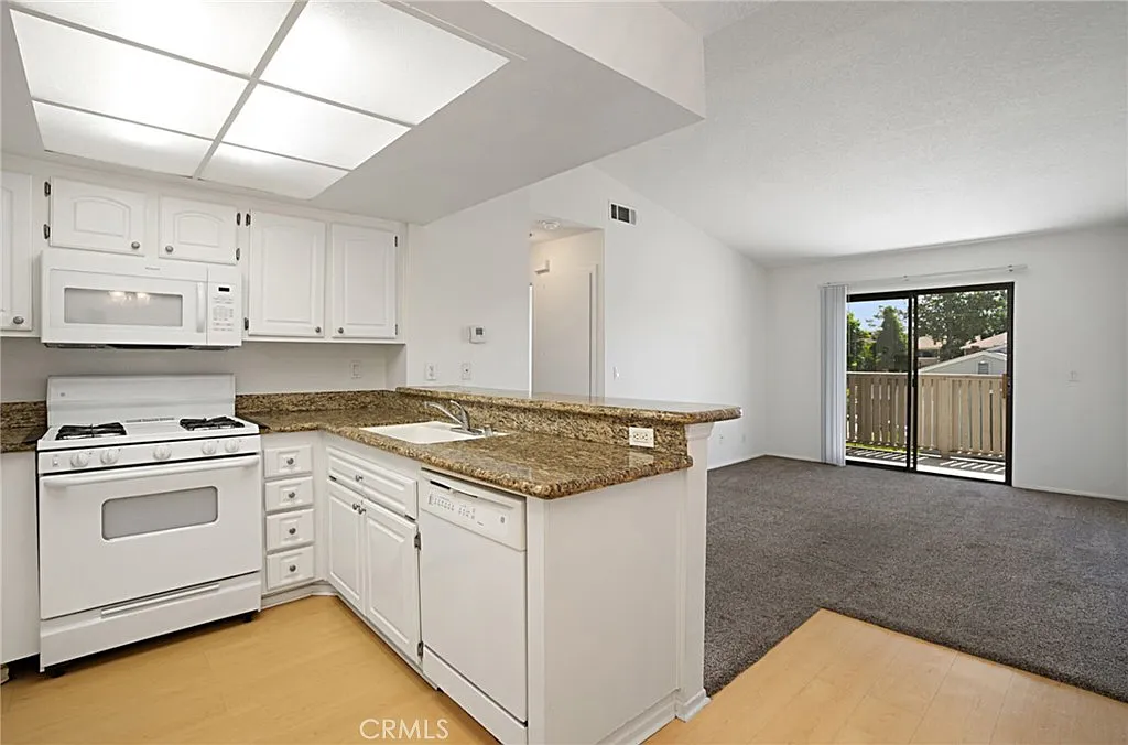 Kitchen with white cabinets, granite countertops, stove, microwave, and sink, with adjacent living area and sliding door to balcony.
