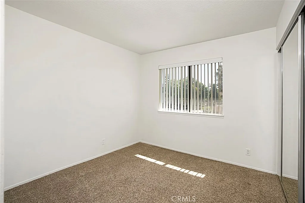 Vacant room with beige carpet, white walls, and window with vertical blinds.
