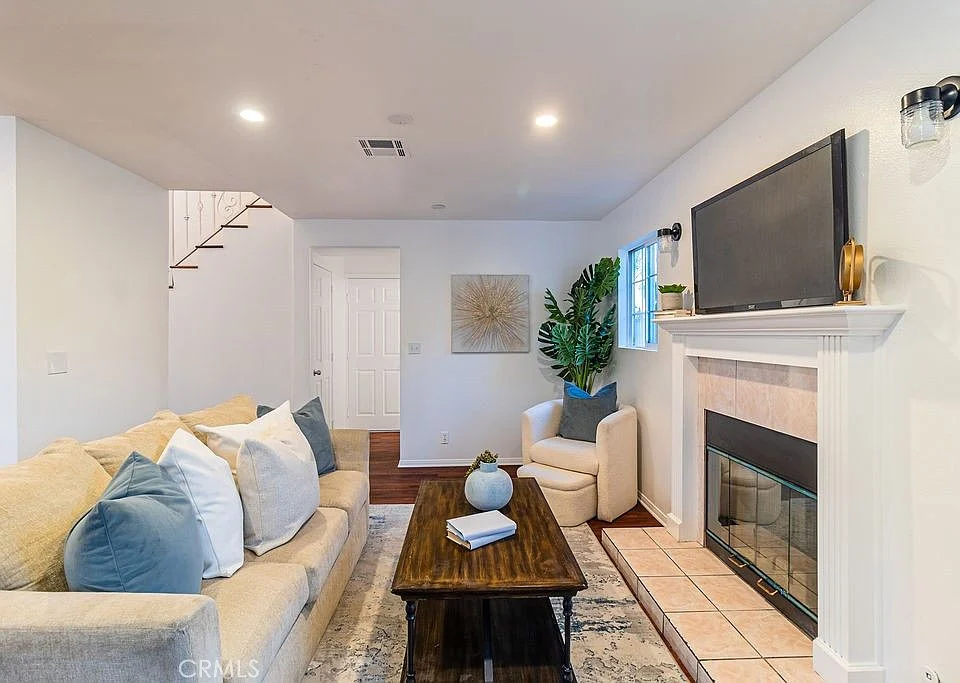living room with beige sofa, blue and white cushions, wooden coffee table, fireplace, TV, plant, artwork, white walls, wooden floor, and staircase