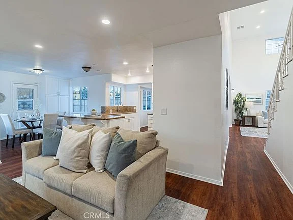 living room with beige couch, decorative pillows, wooden floor, windows, dining area, kitchen, and staircase