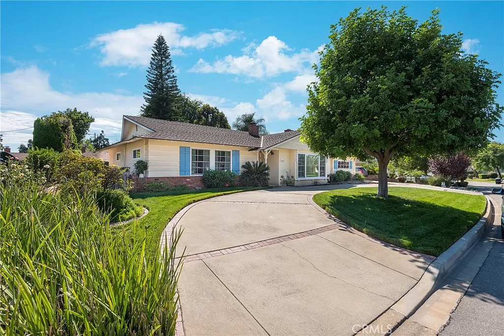 Luxury single-story yellow house with blue shutters, paved driveway, porch, and lush greenery, California real estate