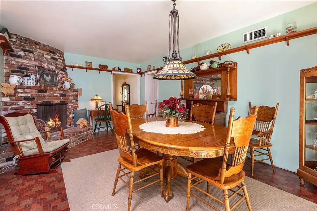 Dining room with round wooden table, six chairs, chandelier, brick fireplace, and decorative shelves