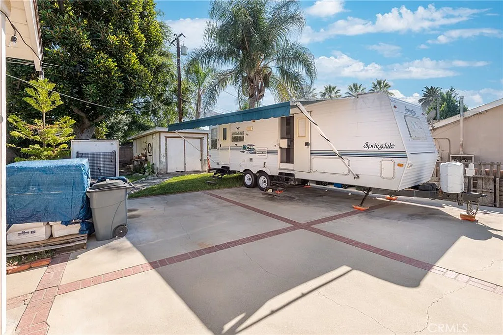 Travel trailer on driveway beside outbuilding, with palm trees and grass around