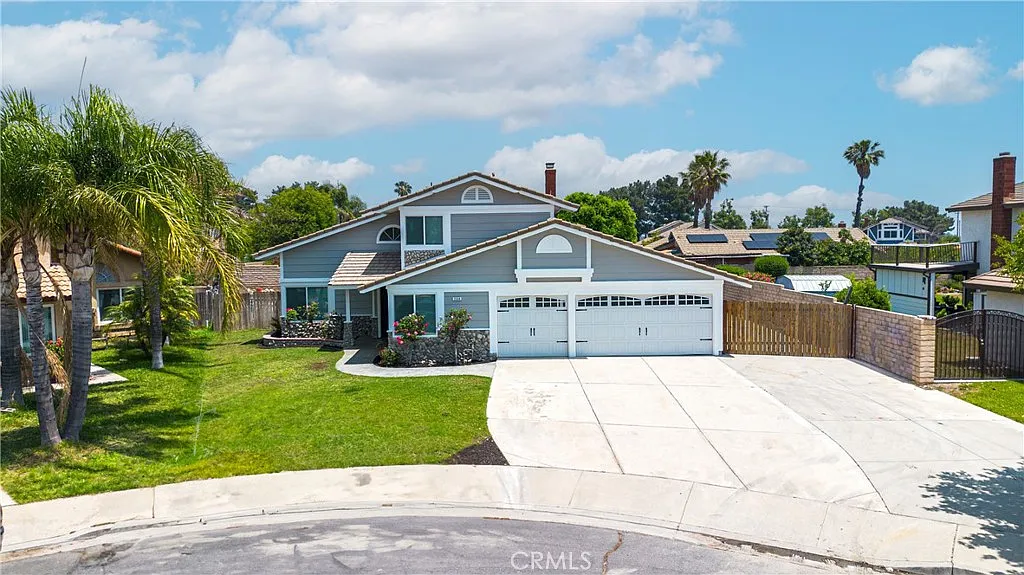 Aerial view of house with circular driveway, palm trees, green lawn, gray exterior, and double garage, real estate.