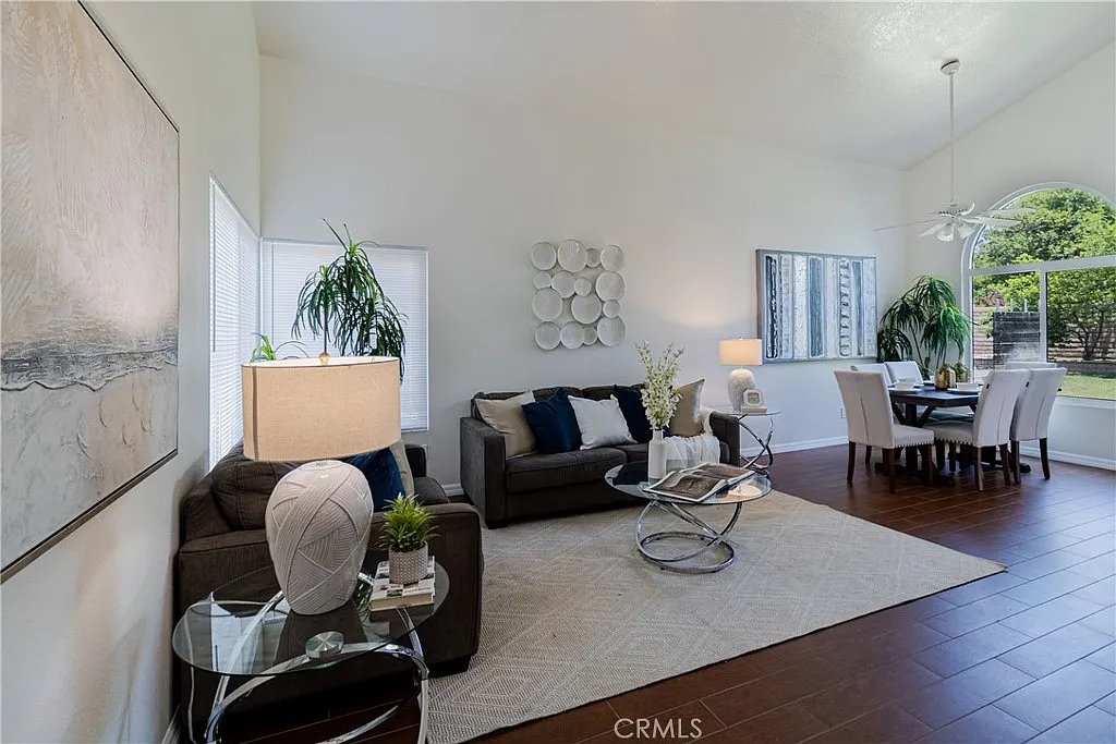 Living room with light sofa, decorative plants, circular coffee table, dining area with white table and chairs, and large windows with natural light.