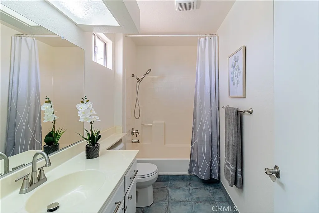 Bathroom with shower/tub, modern vanity with sink, orchid plant, light-colored walls, large mirror, gray shower curtain, and decorative towel.