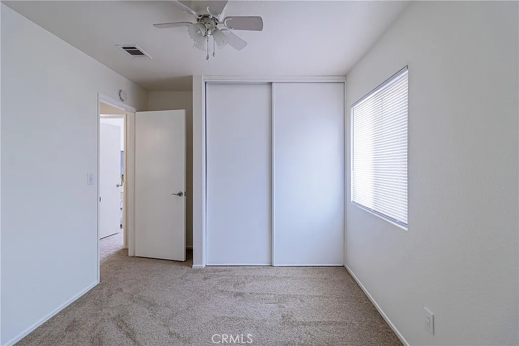 Room with beige carpet, light gray walls, ceiling fan, closet with sliding doors, window with blinds, and open door to another room.