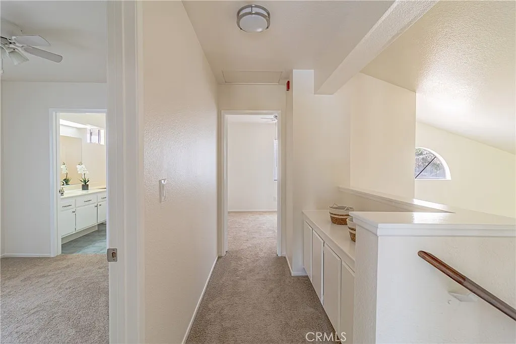 hallway with beige walls and carpet, leading to rooms on both sides, with small arched window at far end.