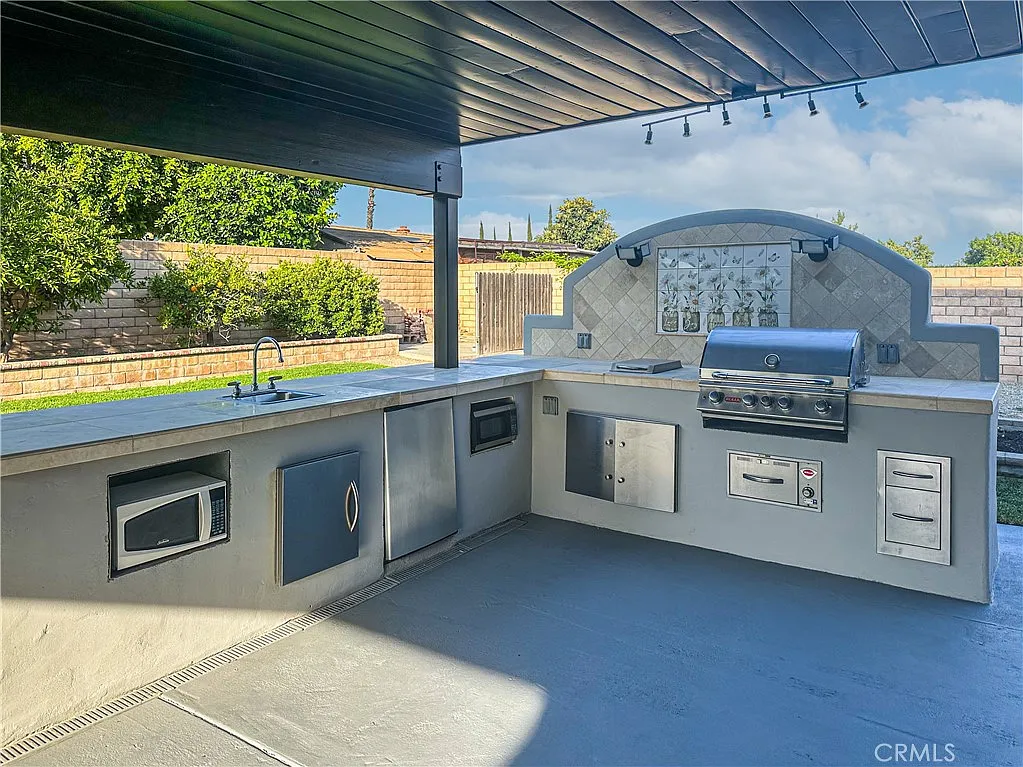 Outdoor kitchen with grill, sink, storage under shaded patio, with trees and fence in background.