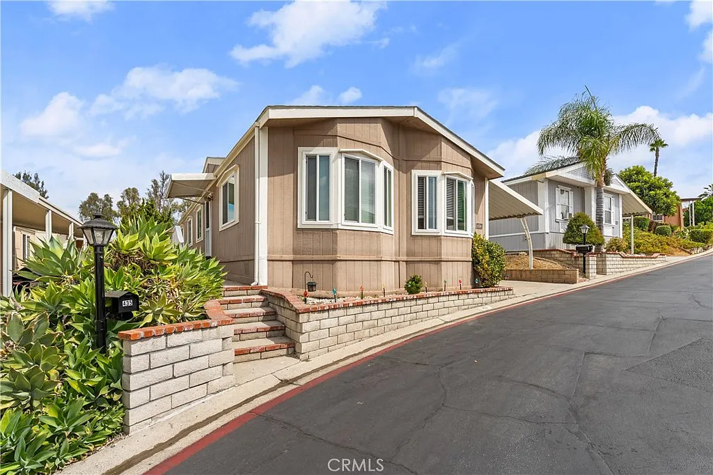 Mobile home California real estate with sloped driveway, palm trees, brick landscaping, and windows.