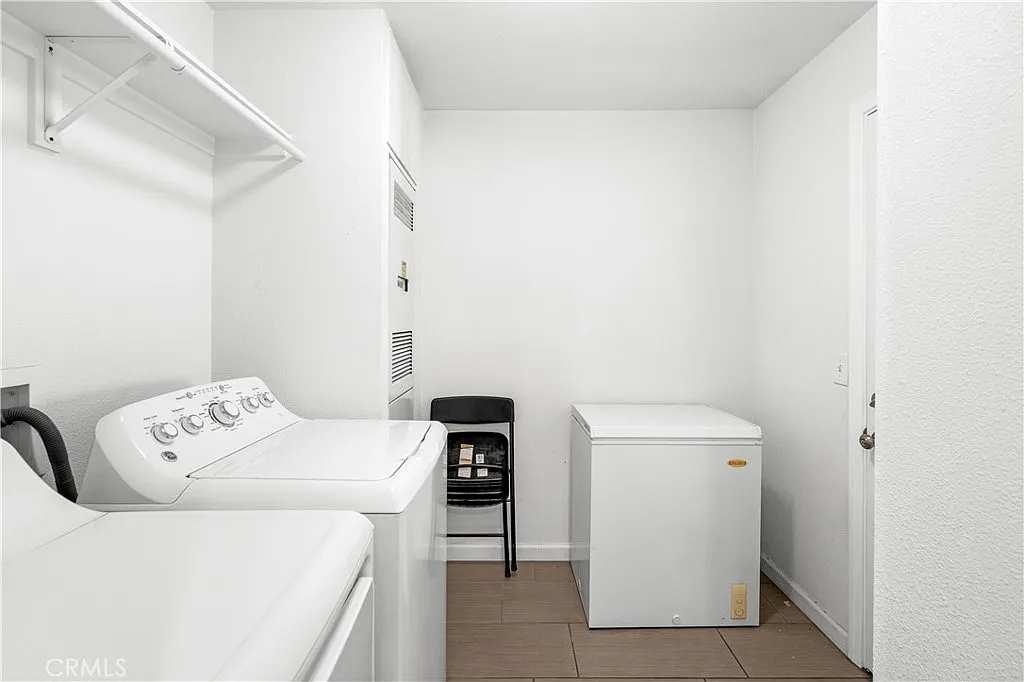 Laundry room with washing machine, dryer, freezer, chair, white walls, and tiled floor.