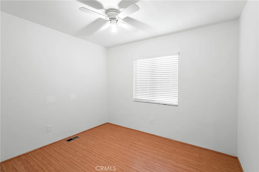 Empty room with ceiling fan, light walls, wooden floor, and window with blinds.