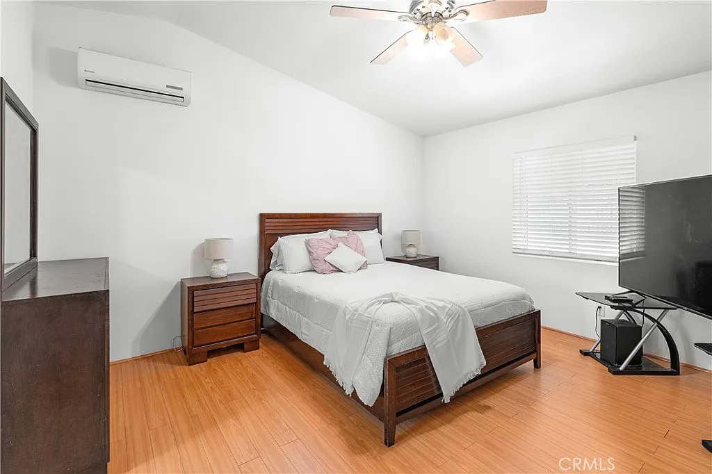 Bedroom with bed, white linens, wooden nightstands and dresser, TV on stand, and wall-mounted air conditioning unit.