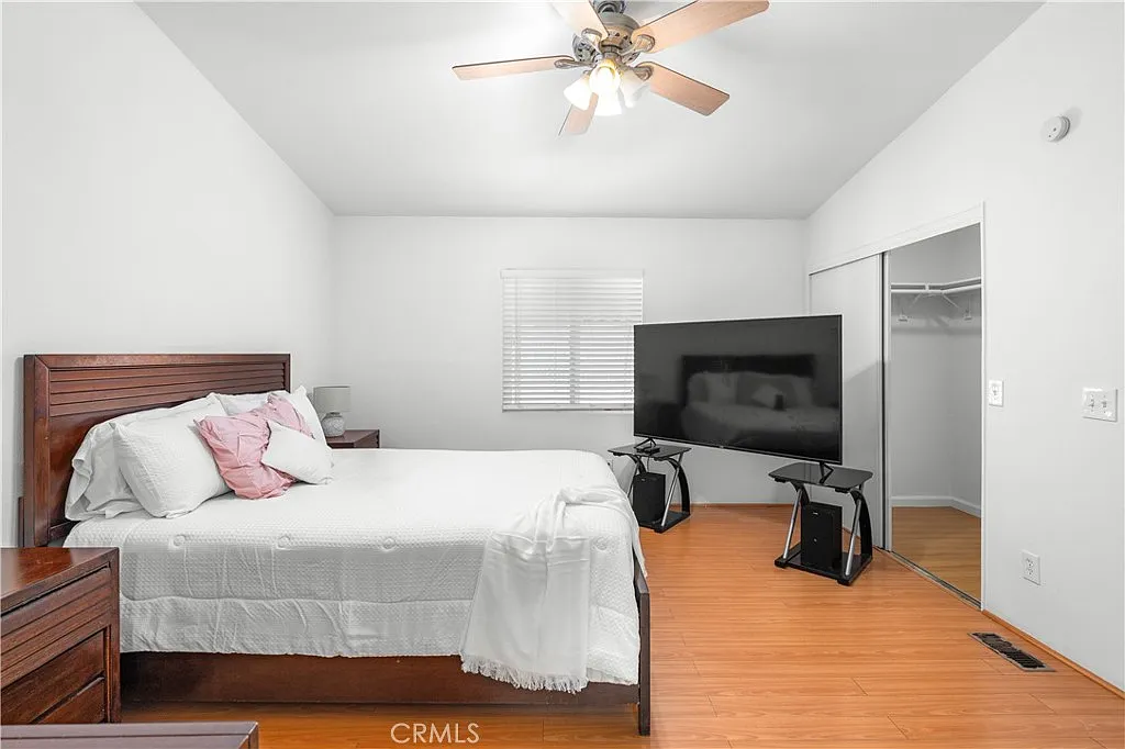 Bedroom with bed, light bedding, ceiling fan, large TV, closet, hardwood floor, and window with blinds.