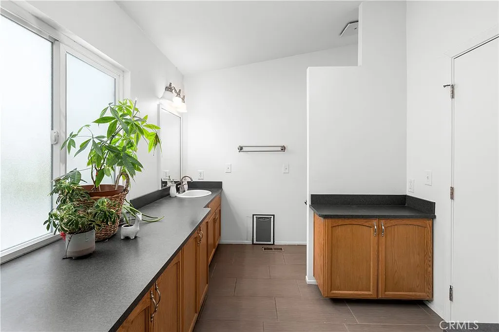 Bathroom with large window, plant, sink, mirror, dark counter, and wood cabinets.