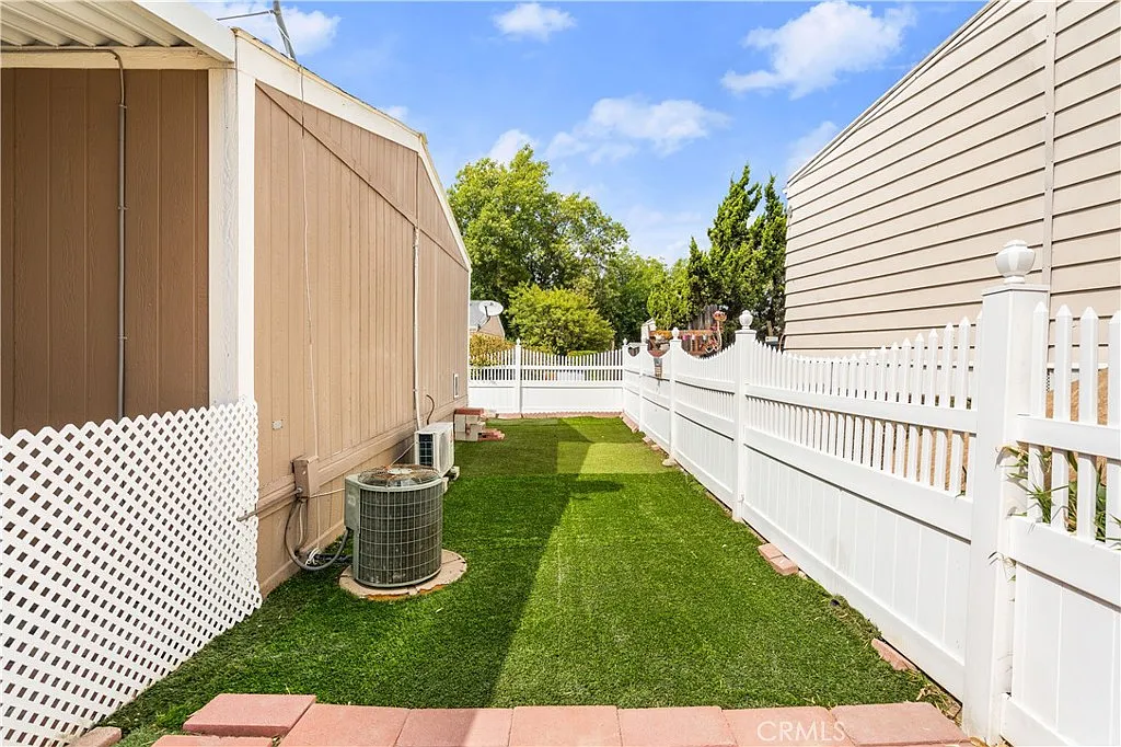 Narrow pathway with artificial grass, white picket fence, and air conditioning unit near building.