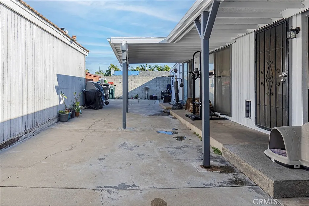 Concrete patio with covered area, potted plants, outdoor pet house, and light-colored buildings on either side.