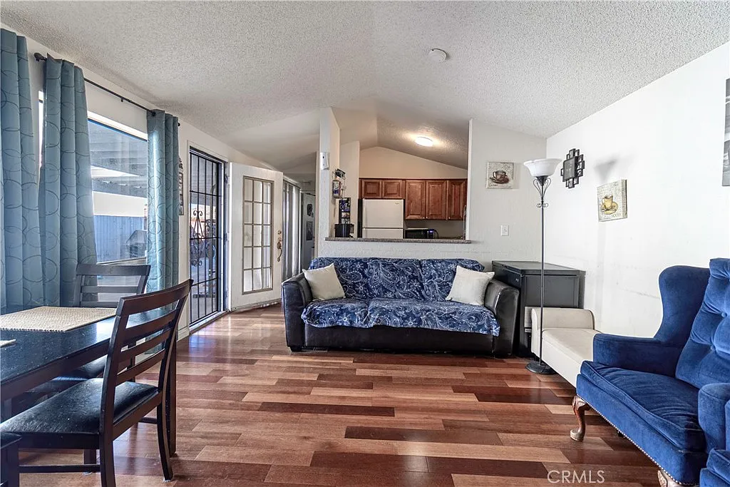 Living room with wooden floors, blue couch and armchair, white walls, large windows with blue curtains, dining table, and adjoining kitchen.